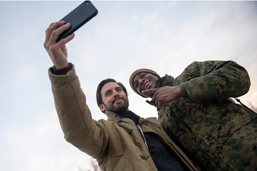 Milo Ventimiglia holds up a phone and poses for a selfie with a smiling service member.