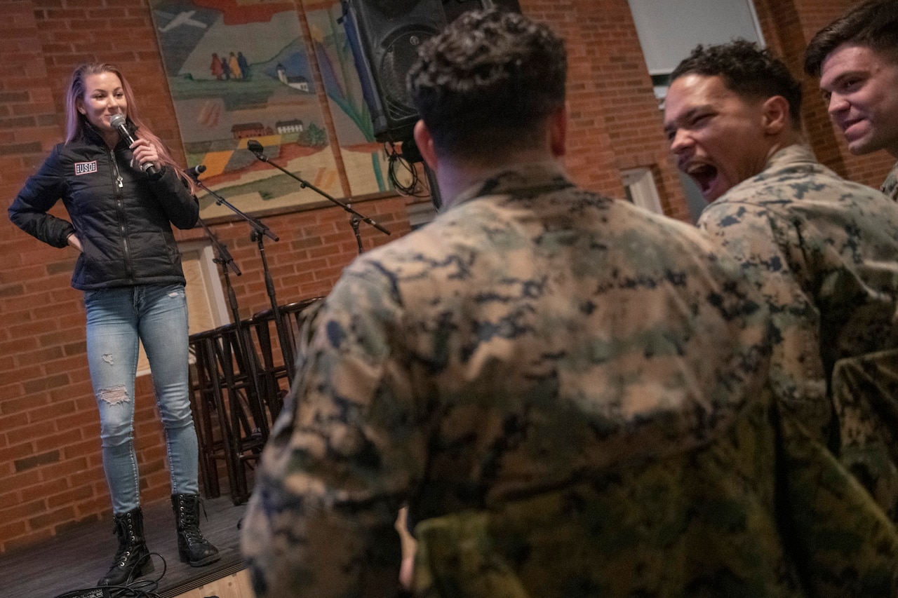 Troops laugh as Comedian Jessiemae Peluso performs on a stage.