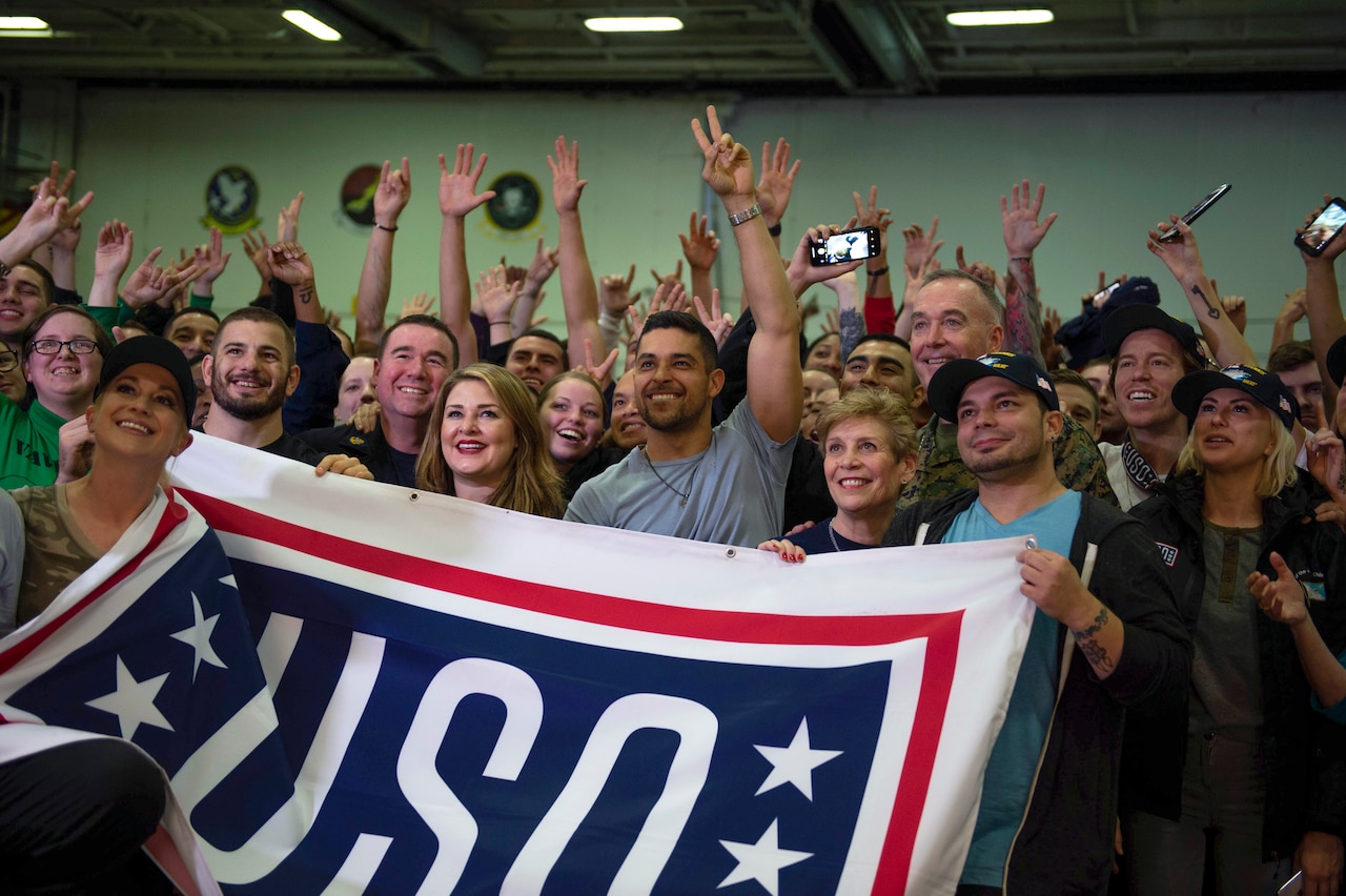 Marine Corps Gen. Joe Dunford, his wife, entertainers and sailors pose for a photo while holding a USO sign.