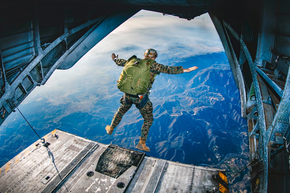 A Marine jumps out of the back of an helicopter flying over mountains.