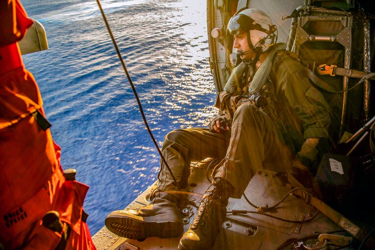 A sailor looks out of the doors of a Seahawk helicopter mid-flight.