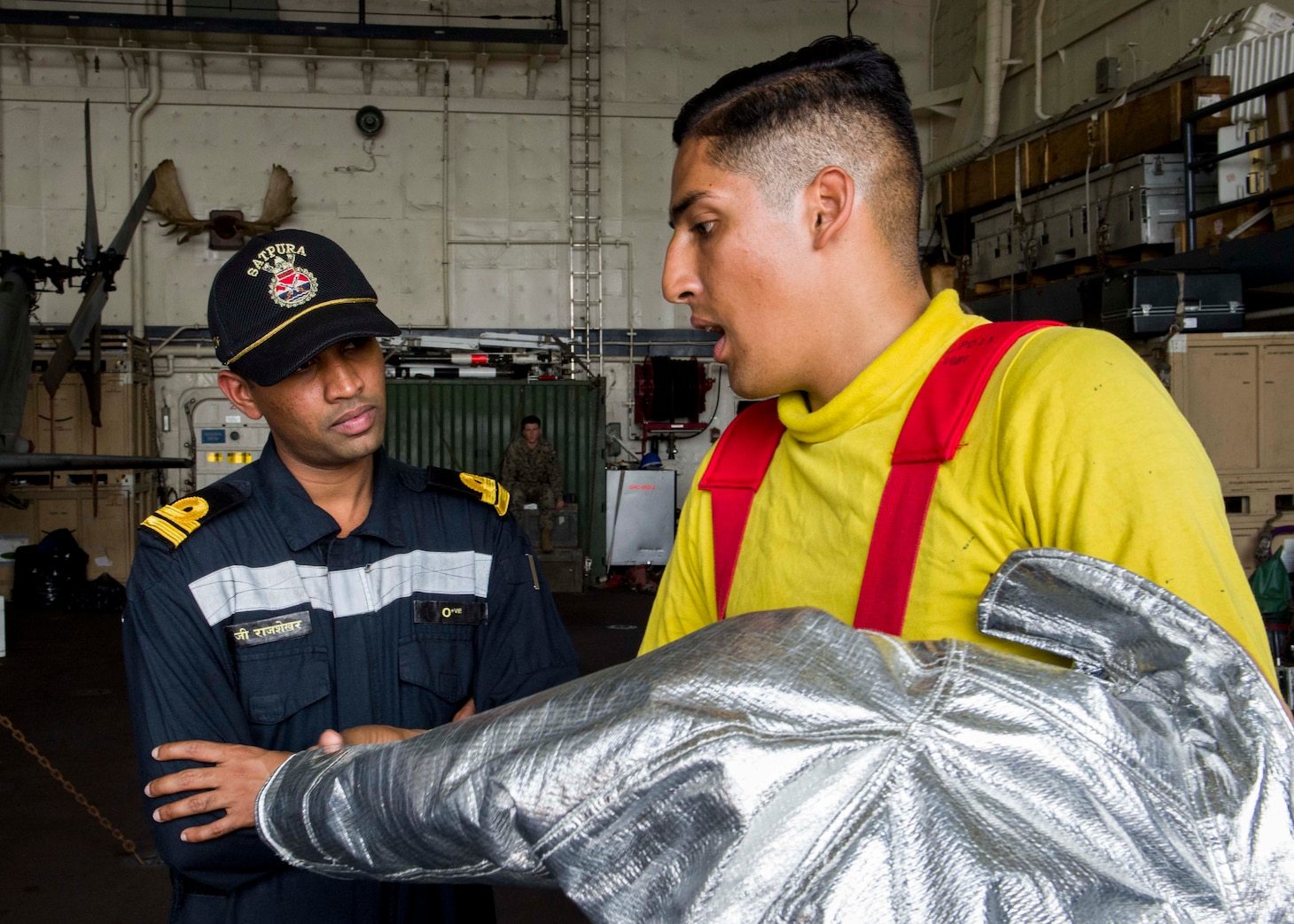 INDIAN OCEAN (Dec. 26, 2018) Aviation Boatswain’s Mate (Handling) 3rd Class Frederick Medina, right, demonstrates the fire protection suit to a Indian naval officer during a tour aboard the San Antonio-class amphibious transport dock ship USS Anchorage (LPD 23) while conducting a cooperative deployment with the Indian navy. Anchorage is deployed as part of the Essex Amphibious Ready Group (ARG) and 13th Marine Expeditionary Unit (MEU).