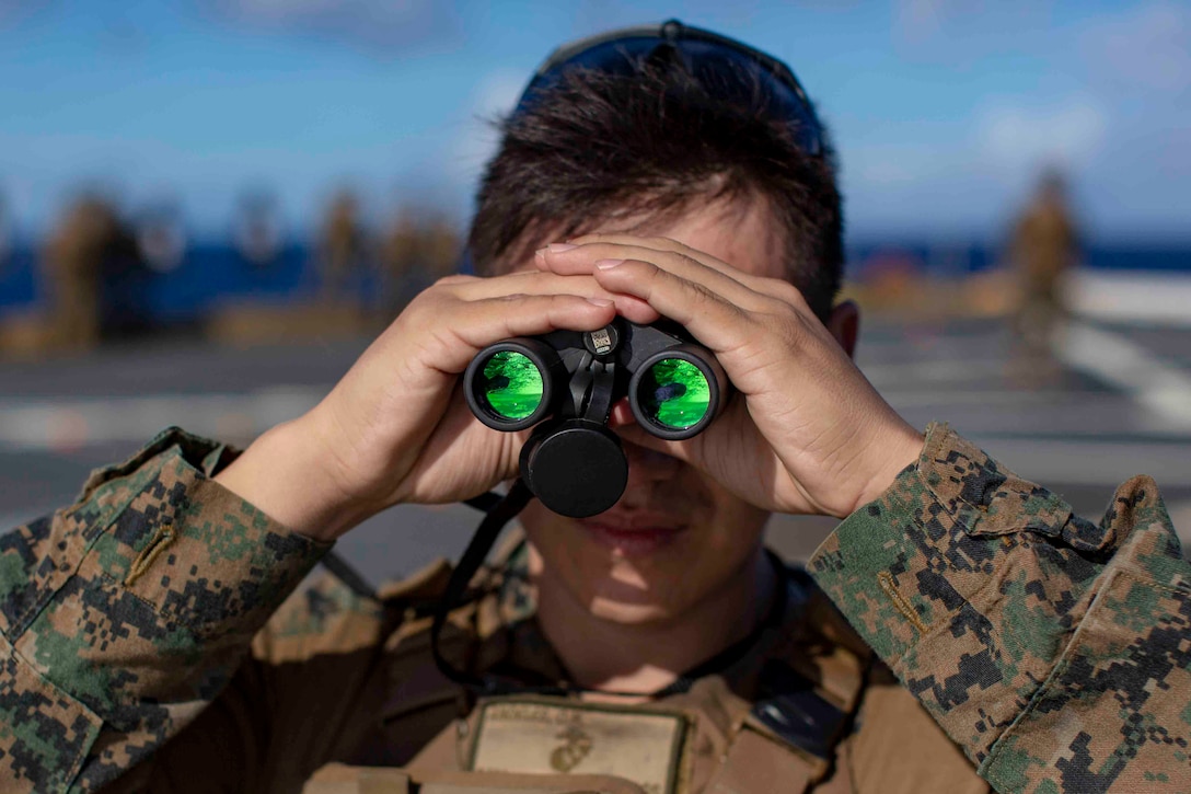 A Marine uses binoculars to watch for hazards while training on a military vessel.