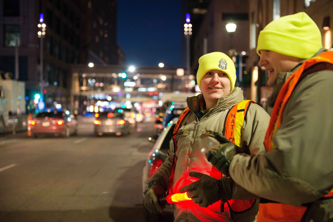 Two guardsmen wearing reflective vests stand on the side of a road.