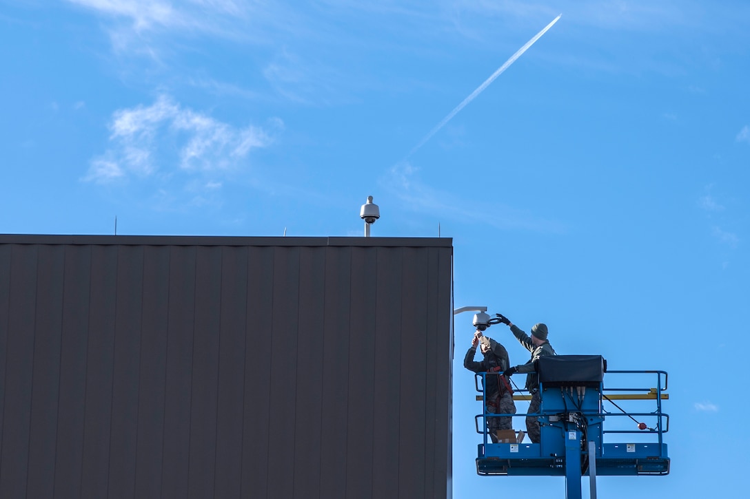 Two airmen work on a security camera at the top of a building.