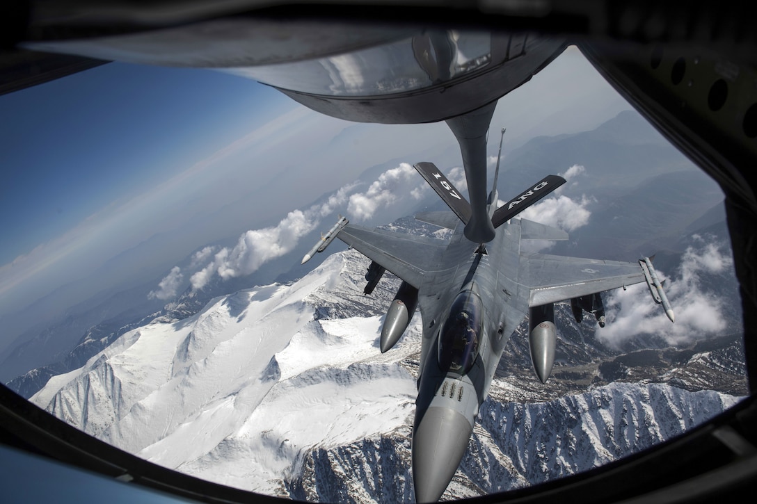 A jet receives fuel from the boom of another aircraft while in flight over snowy mountain peaks.