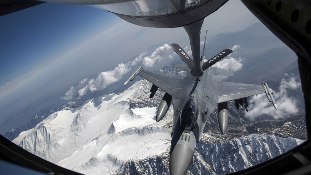 A jet receives fuel from the boom of another aircraft while in flight over snowy mountain peaks.