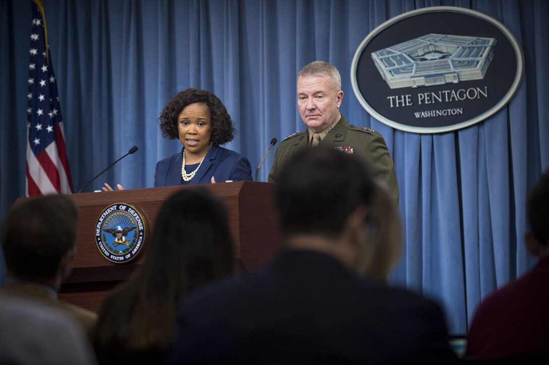 A civilian speaks to reporters while standing with a Marine behind a lectern.