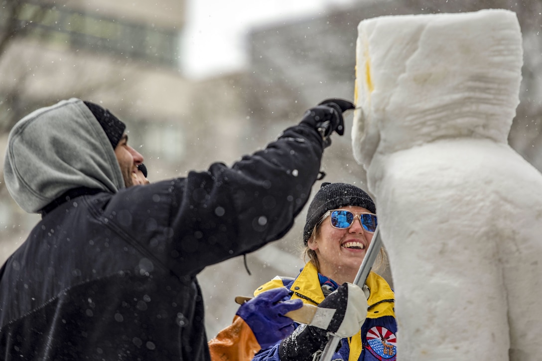 Two sailors add details to a tall snow sculpture during a festival.