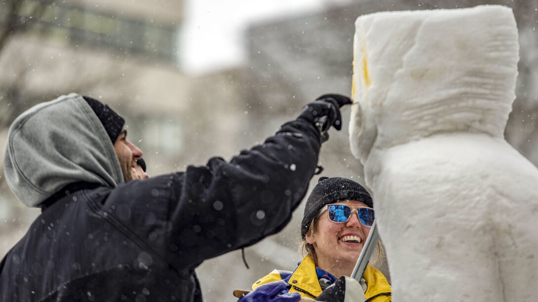 Two sailors add details to a tall snow sculpture during a festival.