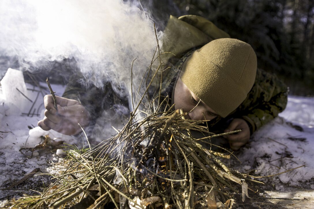 A sailor blows on a fire in kindling wood in the snow.