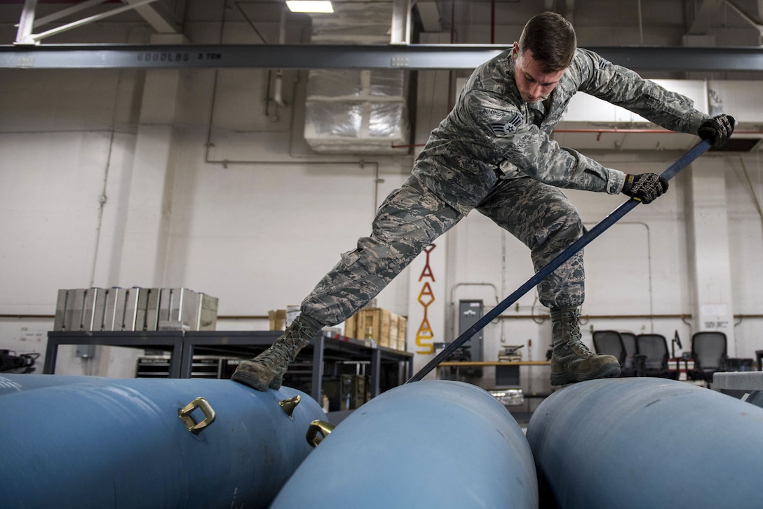 An airman uses a long bar to roll one of three blue bomb bodies.