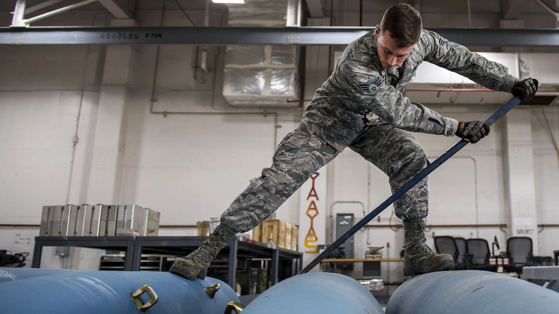 An airman uses a long bar to roll one of three blue bomb bodies.