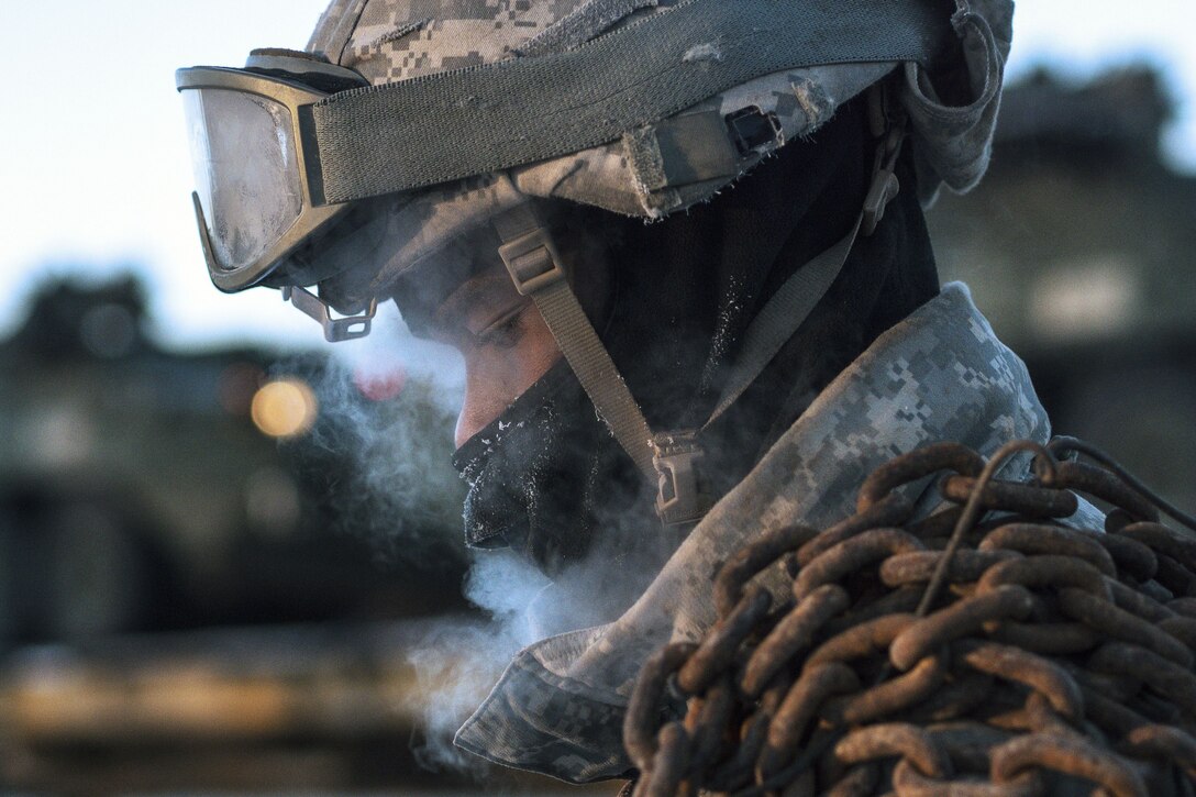 Fog from the cold wafts around the face of a soldier wearing a balaclava with a chain slung over his shoulder.