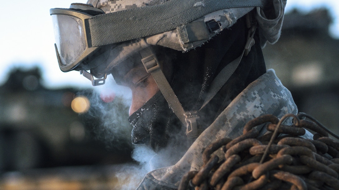 Fog from the cold wafts around the face of a soldier wearing a balaclava with a chain slung over his shoulder.