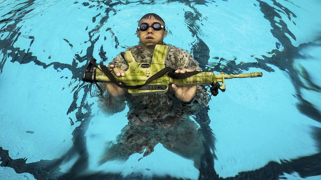 A Marine holds a yellow rubber rifle while he treads water in a pool.