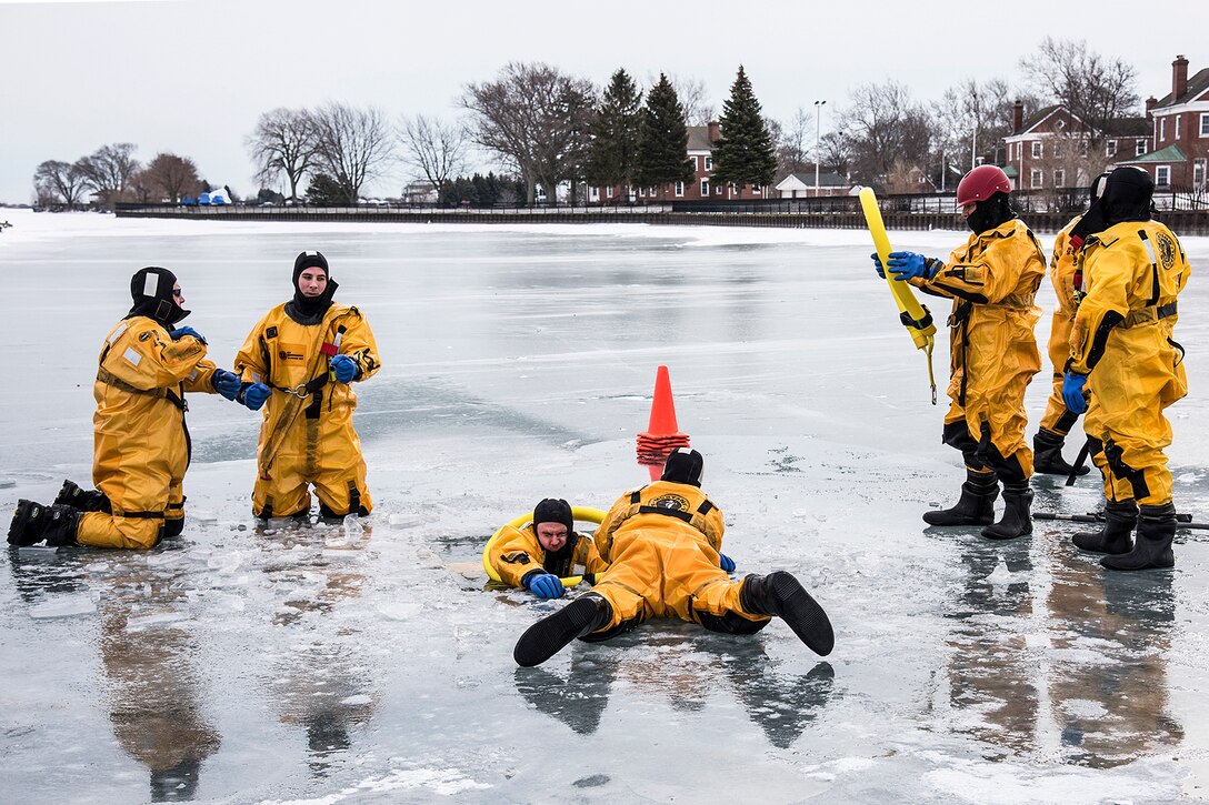 Firefighters in yellow gear stand on a frozen lake while pulling a person from a hole in the ice.