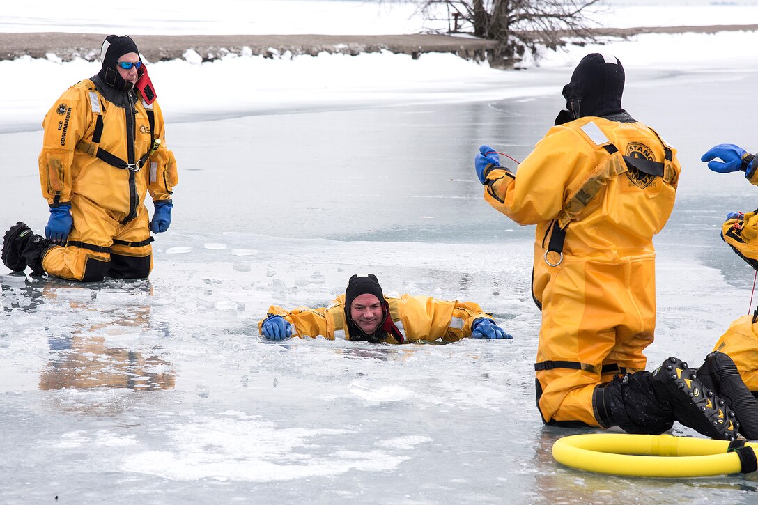 Two firefighters kneel on a frozen lake while a third is submerged in a hole in the ice.