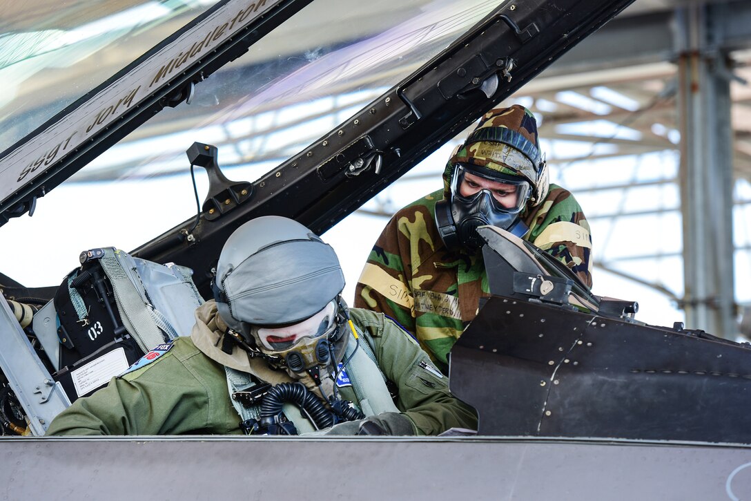 A Guardsman wearing a chemical suit assists a pilot into the cockpit of an aircraft.