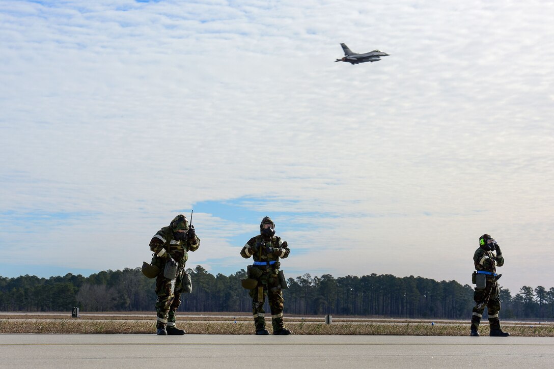 Three guardsmen wearing chemical suits observe aircraft taking off on the flightline.