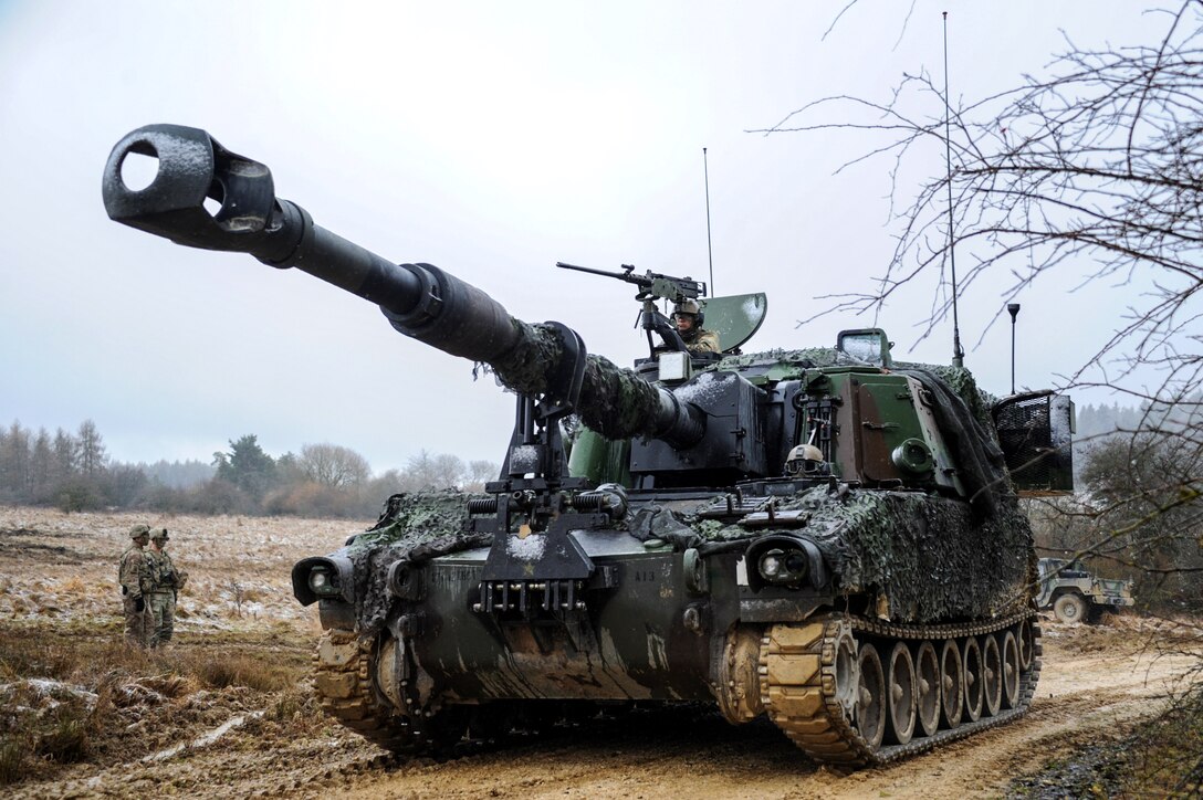 Two soldiers observe from the side of a dirt road as team members maneuver a M109A6 Paladin howitzer.