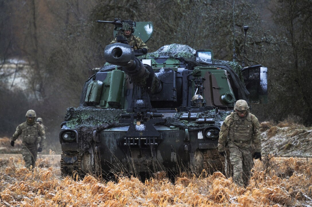 Soldiers walk along and in front of an M109A6 Paladin howitzer.