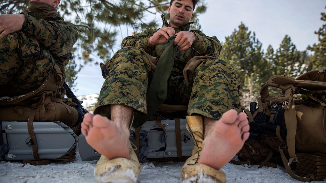 A Marine perches his bare feet on his boots while sitting on gear and changing his socks in the snow.