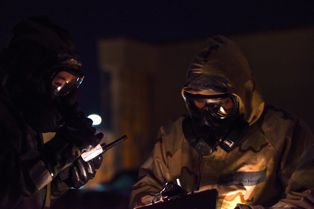 Two airmen in protective suits use a flashlight to read from a clipboard.
