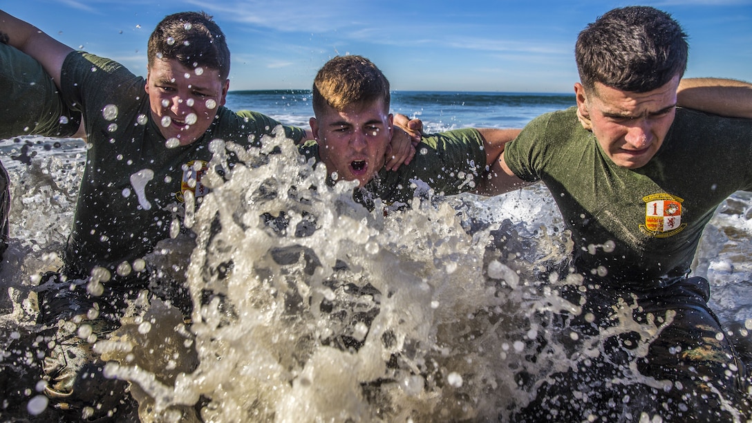 Surf splashes up as three Marines with their arms around each other's shoulders move through waist-deep water at the beach.