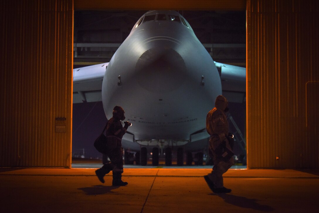 Two airmen walk in a darkened hangar past an opening that reveals the nose of large parked aircraft.