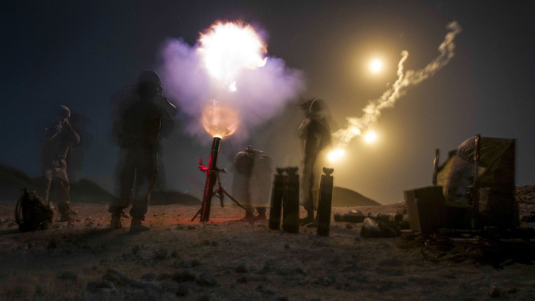 A blast from a mortar lights up the night sky, as soldiers stand by the weapon.