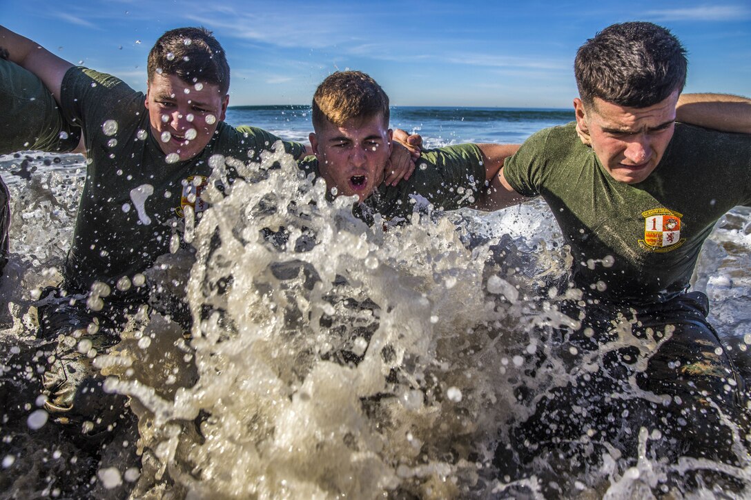 Surf splashes up as three Marines with their arms around each other's shoulders move through waist-deep water at the beach.