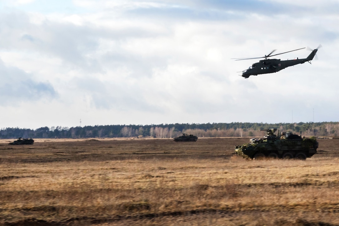 A Polish helicopter flies over Stryker vehicles driving through an open field.