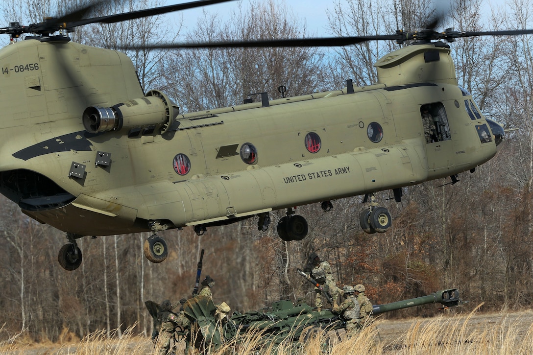 Soldiers prepare to hook up a howitzer to a hovering helicopter.