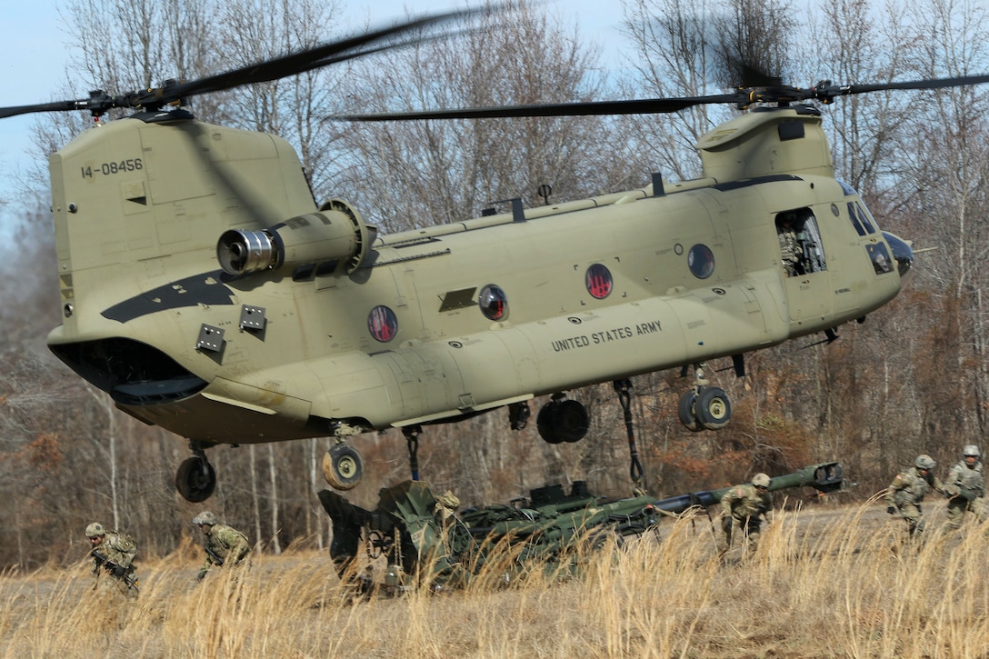 Soldiers run from a hovering helicopter connected to a howitzer.