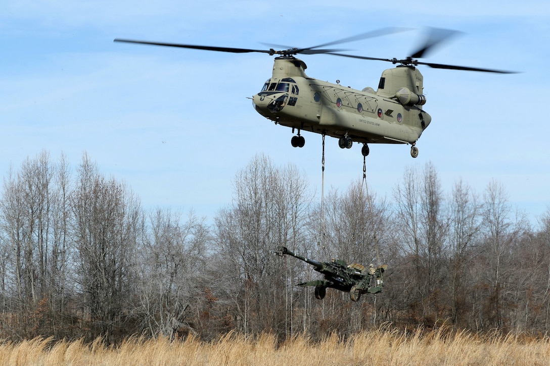 A helicopter lifts off carrying a howitzer hooked from its bottom.