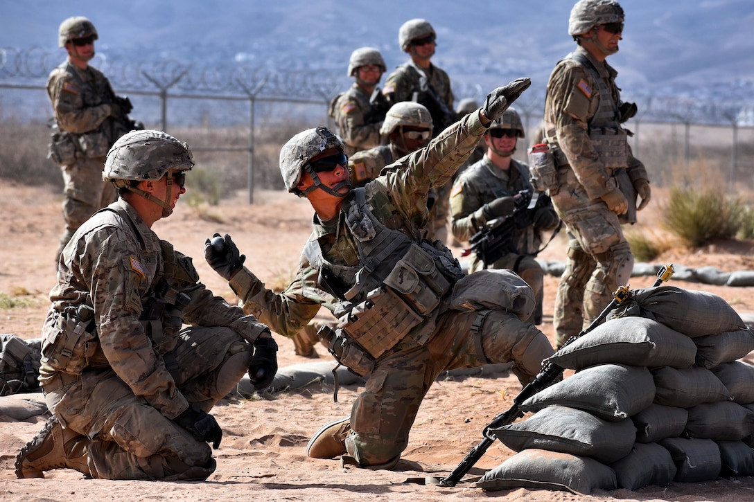 A soldier throws a training grenade from behind a pile of sandbags.