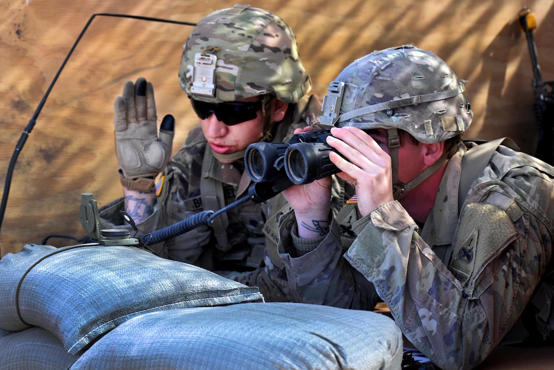 Two soldiers crouch behind a pile of sandbags while one of them looks through binoculars.