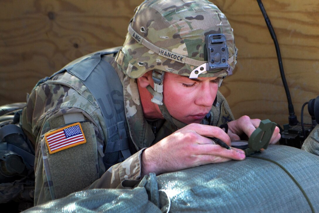 A soldier looks at a lensatic compass while leaning on a sandbag.