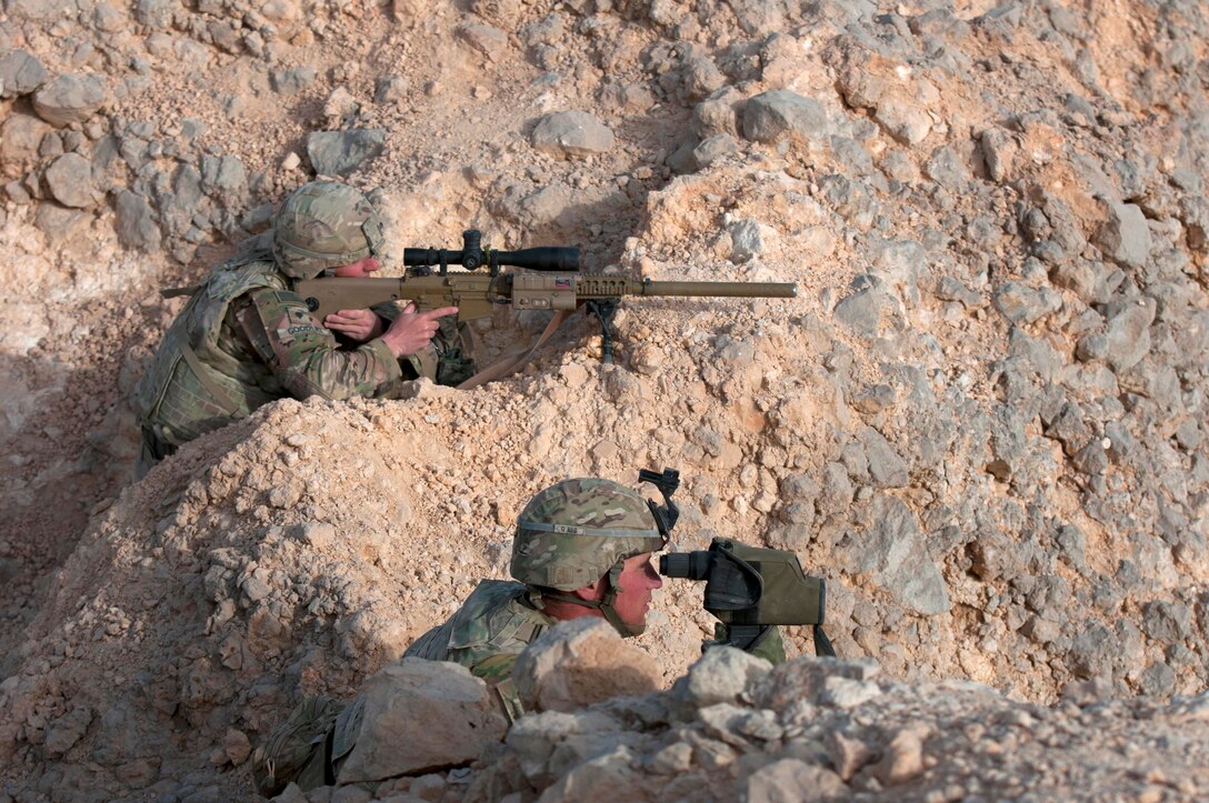 Two soldiers scan an area while taking cover in rocky terrain.