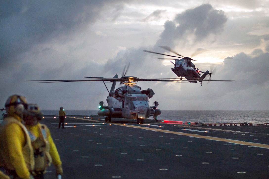 A CH-53E Super Stallion helicopter prepares to take off while another Super Stallion prepares to land on a flight deck.