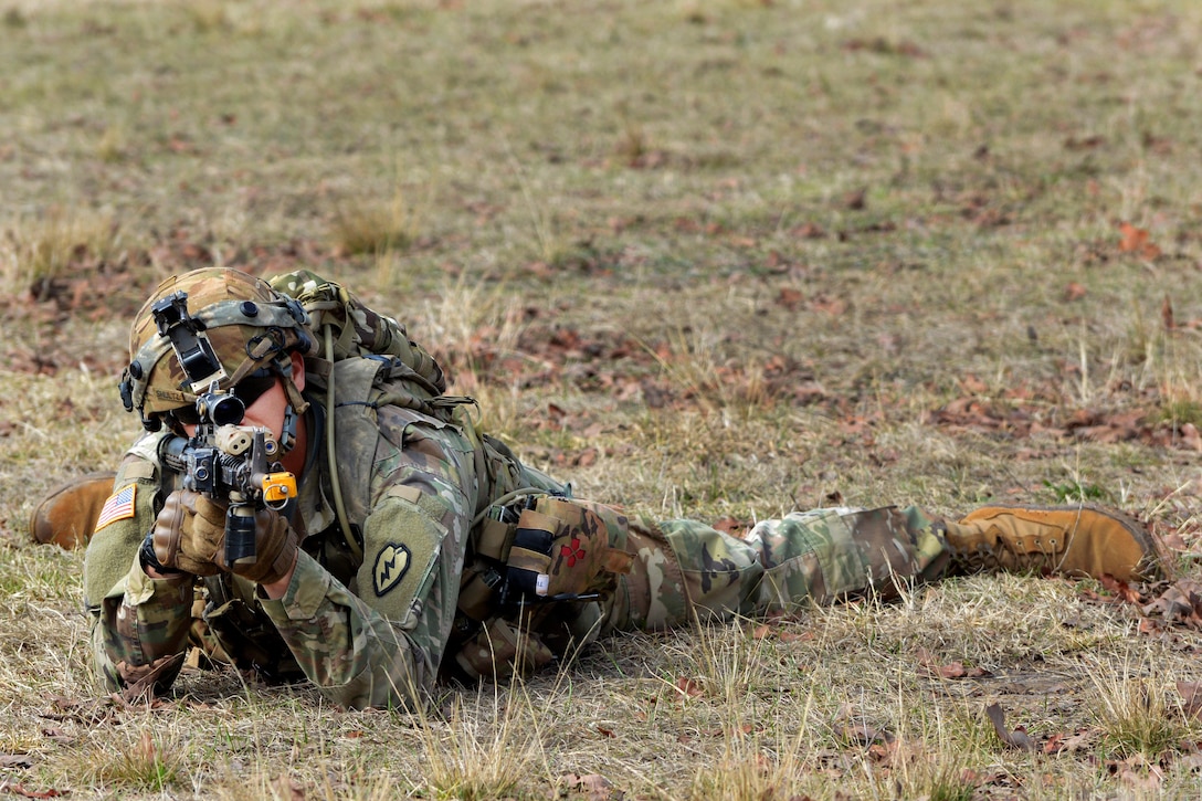 A soldier looks through a rifle scope while laying on the ground.
