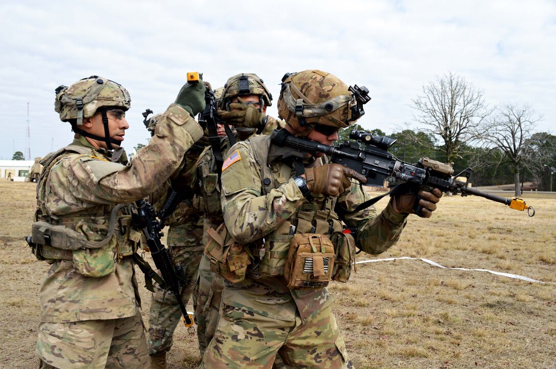 A soldier helps position another soldier's rifle while a third soldier looks through a rifle scope.