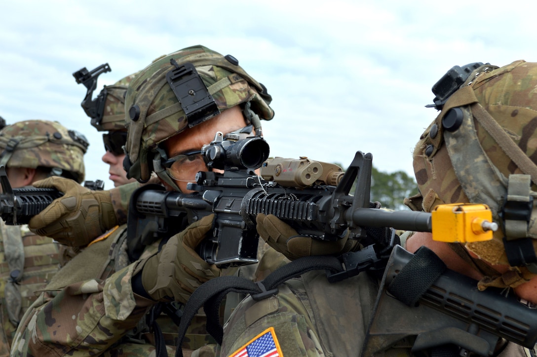 A soldier looks through a rifle scope while the weapon is propped on another soldier's shoulder.