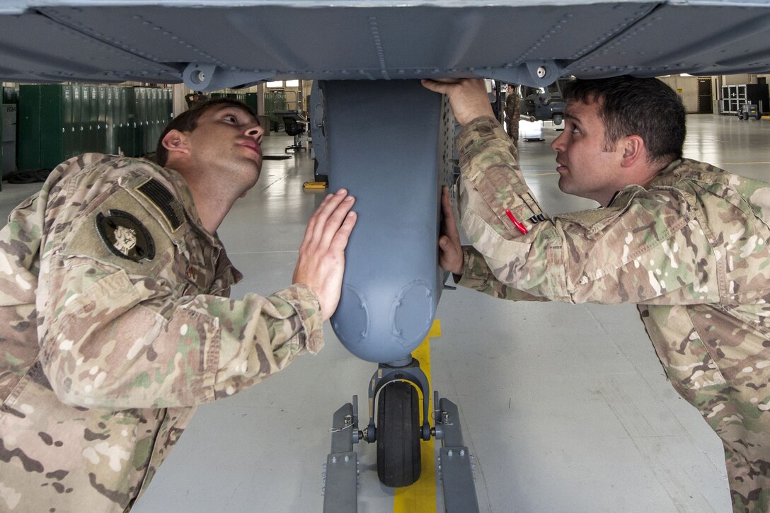 Two airmen examine the horizontal stabilizer on the bottom of a helicopter.