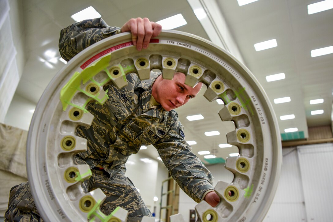 An airman scrutinizes a freestanding wheel while manually rotating it in a well-lit room.