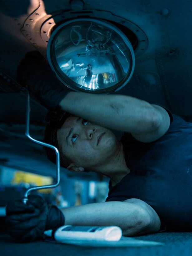 A sailor lays on the ground to conduct maintenance work on a helicopter.