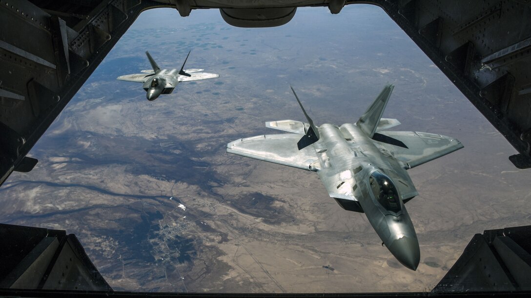 Two fighter jets, viewed from an opening in a third aircraft in front of them, fly over brownish terrain.