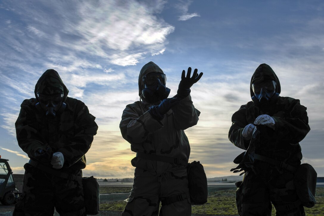 Three airmen, shown in silhouette, put on protective gear while standing in a row outside.