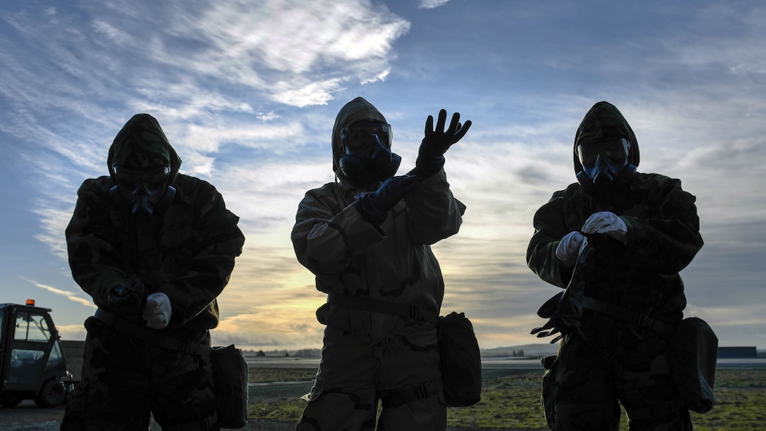 Three airmen, shown in silhouette, put on protective gear while standing in a row outside.